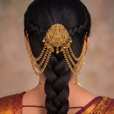 A close-up back view of a woman wearing an ornate gold hair accessory with dangling chains and a central decorative piece, styled in a long braid. The background is a soft beige studio backdrop.