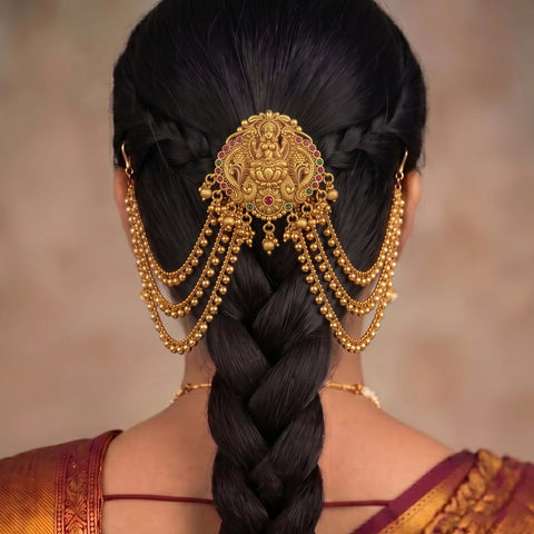 A close-up back view of a woman wearing an ornate gold hair accessory with dangling chains and a central decorative piece, styled in a long braid. The background is a soft beige studio backdrop.