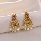 A pair of ornate gold-toned chandelier earrings with white faux pearls, small green and red stones, and clear rhinestones, displayed on a soft white fabric surface.