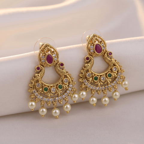 A pair of ornate gold-toned traditional Indian chandbali earrings with red, green, and white stones, displayed on a soft white fabric background.