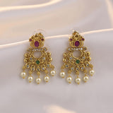 A pair of gold-toned traditional Indian chandelier earrings with white faux pearls, green and red stones, and clear rhinestones, displayed on a draped white satin fabric.