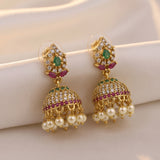 A pair of gold-plated Indian jhumka earrings adorned with green teardrop crystals, pink gemstones, clear pavé stones, and dangling white faux pearls, displayed on a soft cream satin fabric.