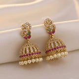 A pair of gold-plated Indian jhumka earrings adorned with clear crystals, small pinkish-red stones, and white faux pearls, displayed on a soft beige fabric background.