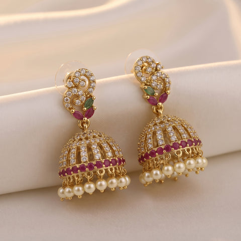 A pair of gold-plated Indian jhumka earrings adorned with clear crystals, small pinkish-red stones, and white faux pearls, displayed on a soft beige fabric background.