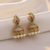 A pair of gold-plated Indian jhumka earrings adorned with clear pavé stones, pink gemstones, and dangling white pearls, displayed on a soft cream satin fabric.