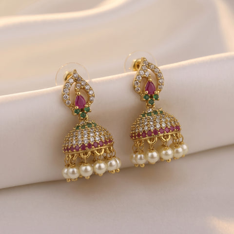 A pair of gold-plated Indian jhumka earrings adorned with clear pavé stones, pink gemstones, and dangling white pearls, displayed on a soft cream satin fabric.