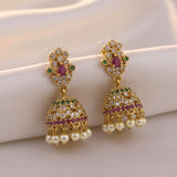 A pair of gold-plated Indian jhumka earrings decorated with clear crystals, green and pink gemstones, and white faux pearls, displayed on a soft beige fabric background.