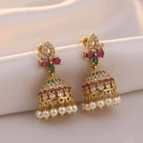 A pair of gold-plated Indian jhumka earrings decorated with green and pink stones, clear crystals, and white faux pearls, displayed on a soft beige fabric background.