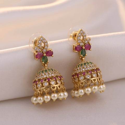 A pair of gold-plated Indian jhumka earrings decorated with green and pink stones, clear crystals, and white faux pearls, displayed on a soft beige fabric background.