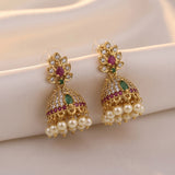 A pair of gold-plated Indian jhumka earrings decorated with green and red stones, clear crystals, and white faux pearls, displayed on a soft beige fabric background.