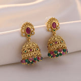 A pair of ornate gold-toned Indian jhumka earrings with oval pinkish-red stones, small green stones, and dangling clusters of pink and green faceted beads, displayed on a soft white fabric surface.