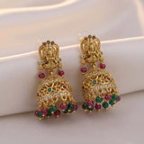 A pair of ornate gold-toned Indian jhumka earrings with detailed filigree, small clear stones, and dangling red and green gemstone accents, displayed on a soft white fabric surface.