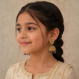 A young girl with dark hair in a braid wearing a gold-toned traditional jhumka earring with red and green stone accents, set against a soft beige background.