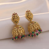 A pair of ornate gold jhumka earrings with colorful gemstone accents, displayed on a draped cream satin fabric background.