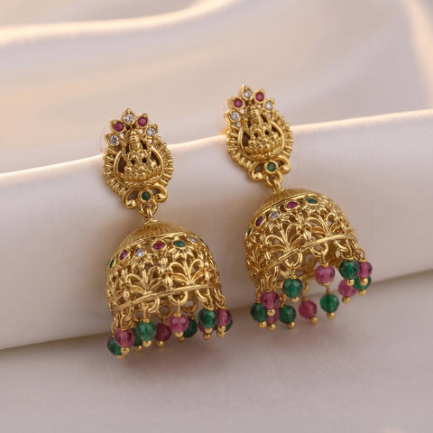 A pair of ornate gold jhumka earrings with colorful gemstone accents, displayed on a draped cream satin fabric background.