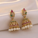 A pair of gold-plated Indian jhumka earrings decorated with pink and green stones, clear crystals, and dangling white faux pearls, displayed on a soft beige fabric background.