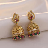 A pair of ornate gold-toned Indian jhumka earrings with red and green stone accents, displayed on a soft cream fabric background.