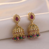 A pair of gold-plated Indian jhumka earrings with teardrop-shaped pinkish-red stones, clear rhinestones, and dangling green and pink bead accents, displayed on a soft white satin fabric.