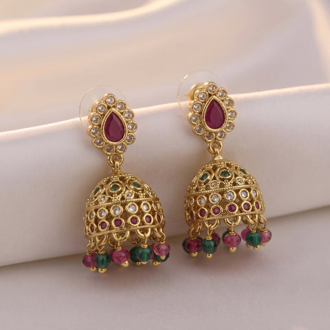 A pair of gold-plated Indian jhumka earrings with teardrop-shaped pinkish-red stones, clear rhinestones, and dangling green and pink bead accents, displayed on a soft white satin fabric.