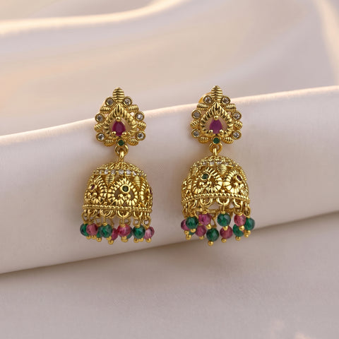A pair of ornate gold-toned Indian jhumka earrings with red and green stone accents, displayed on a soft white fabric background.