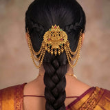 A close-up back view of a woman wearing an ornate gold hair ornament with hanging chains and small red and green stone accents, styled with a long dark braid and sari.