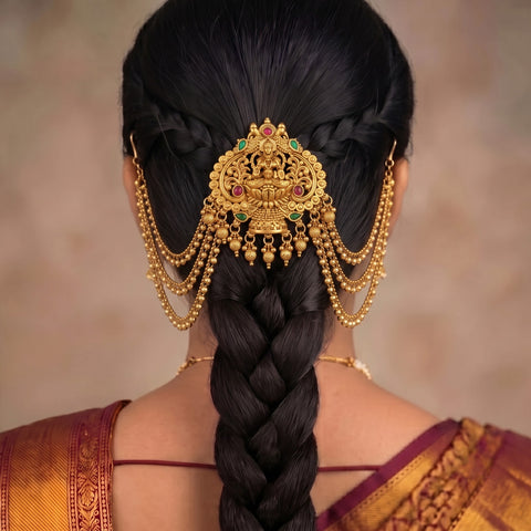A close-up back view of a woman wearing an ornate gold hair ornament with hanging chains and small red and green stone accents, styled with a long dark braid and sari.