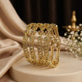A set of ornate gold bangles with green and pink stone accents, displayed on a cream-colored pedestal with draped satin fabric and small white flowers in the background.