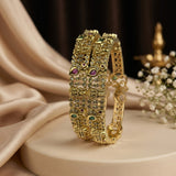 A pair of ornate gold-plated bangles decorated with intricate stone work, featuring small green, red, and white stones, displayed on a beige pedestal with draped satin fabric and baby's breath flowers.