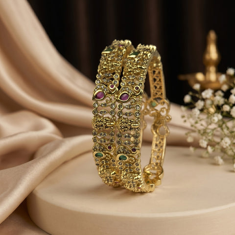 A pair of ornate gold-plated bangles decorated with intricate stone work, featuring small green, red, and white stones, displayed on a beige pedestal with draped satin fabric and baby's breath flowers.