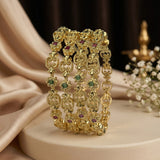 A stack of ornate gold-toned bangles with small green and red gemstone accents, displayed on a cream pedestal with draped satin fabric and white flowers in the background.
