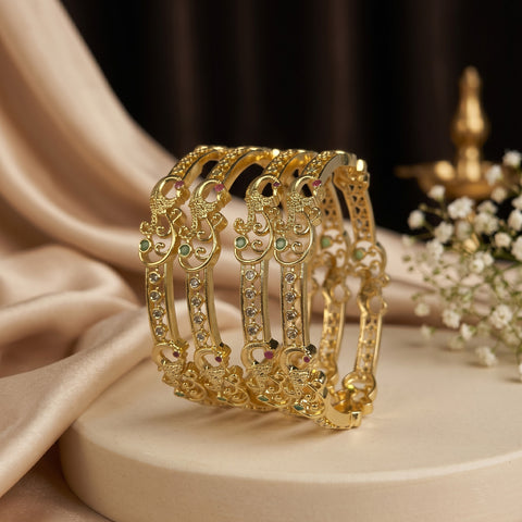 A set of four ornate gold bangles with intricate filigree patterns and small green and pink gemstone accents, displayed on a cream-colored pedestal with draped satin fabric and baby's breath flowers.