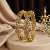 Two ornate gold bangles with green and pink stone accents, displayed on a beige pedestal with draped satin fabric and small white flowers in the background.
