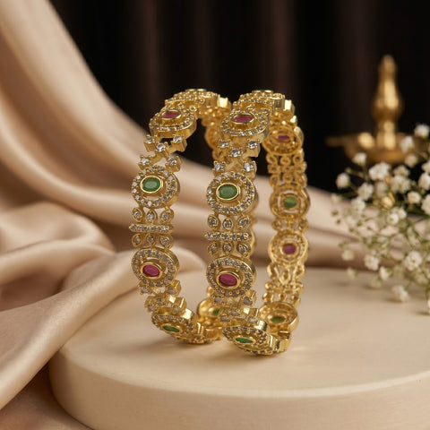 Two ornate gold bangles with green and pink stone accents, displayed on a beige pedestal with draped satin fabric and small white flowers in the background.