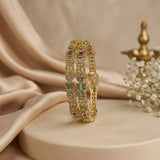 A gold-plated bangle bracelet with green and red stones, displayed on a beige pedestal with draped satin fabric in the background.