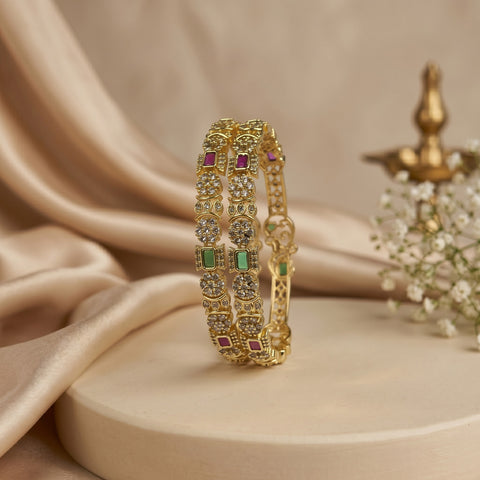 A gold-plated bangle bracelet with green and red stones, displayed on a beige pedestal with draped satin fabric in the background.