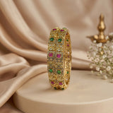 A gold-plated bangle bracelet with green and pink gemstone accents, displayed on a beige pedestal with draped satin fabric in the background.