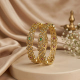 Two ornate gold bangles with green and pink gemstone accents, displayed on a beige pedestal with draped satin fabric and small white flowers in the background.