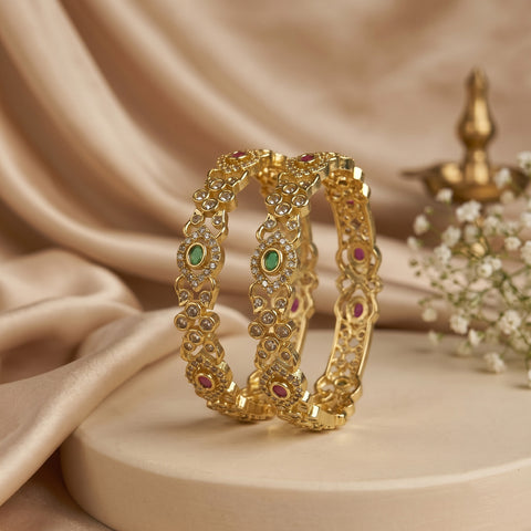 Two ornate gold bangles with green and pink gemstone accents, displayed on a beige pedestal with draped satin fabric and small white flowers in the background.