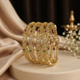 A set of four gold-plated bangles decorated with clear crystals, green and red gemstones, displayed on a cream-colored pedestal with draped satin fabric and baby's breath flowers in the background.