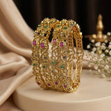 A stack of four ornate gold bangles decorated with green, pink, and clear stones, displayed on a cream-colored pedestal with draped satin fabric and small white flowers in the background.