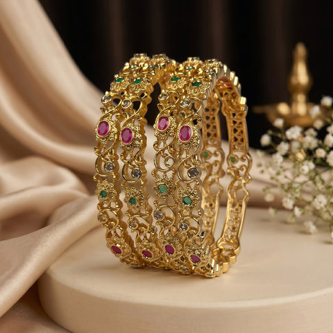 A stack of four ornate gold bangles decorated with green, pink, and clear stones, displayed on a cream-colored pedestal with draped satin fabric and small white flowers in the background.
