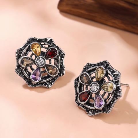 A pair of silver-toned filigree stud earrings with a floral design set with multiple colored gemstones, displayed on a soft peach background.