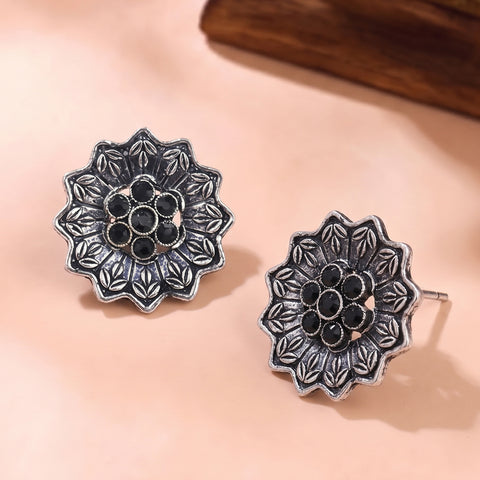 A pair of oxidized silver flower-shaped stud earrings with black circular accents, displayed on a soft peach-colored surface.