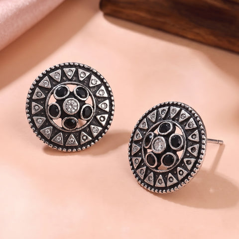 A pair of round silver-toned stud earrings with intricate black and silver geometric patterns and central clear stones, displayed on a soft peach surface.