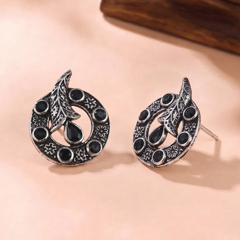 A pair of silver-toned crescent moon stud earrings with black oxidized details, small circular stones, and a teardrop center, displayed on a warm beige surface.