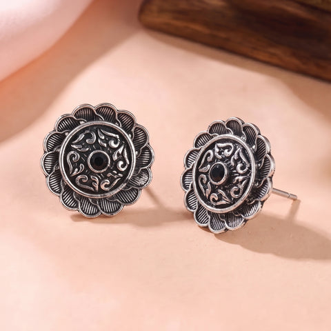 A pair of silver-toned floral stud earrings with intricate filigree detailing and a central black stone, displayed on a soft beige surface.