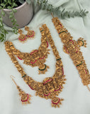 An ornate gold-toned traditional jewelry set arranged on a light fabric background, including a necklace, matching earrings, a bracelet, and a waist chain, decorated with gold filigree, pearl-like beads, and small pink and green accents. A potted plant and greenery are placed around the set as styling props.