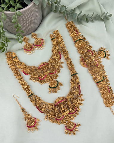 An ornate gold-toned traditional jewelry set arranged on a light fabric background, including a necklace, matching earrings, a bracelet, and a waist chain, decorated with gold filigree, pearl-like beads, and small pink and green accents. A potted plant and greenery are placed around the set as styling props.