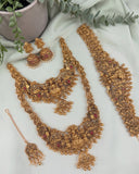 An ornate gold-toned traditional jewelry set arranged on a light fabric background, including a necklace, matching earrings, a bracelet, and a dangling pendant. The pieces are decorated with intricate filigree, small beads, and red stone accents, with green leaves and a textured vase in the background.