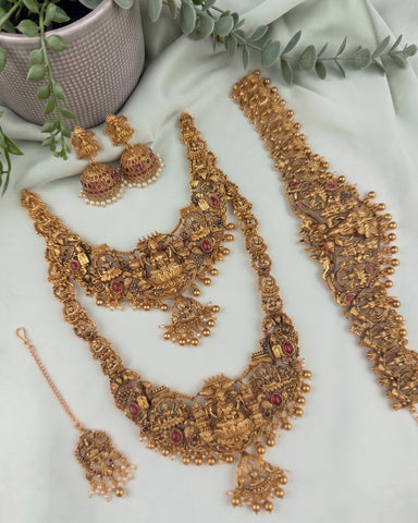 An ornate gold-toned traditional jewelry set arranged on a light fabric background, including a necklace, matching earrings, a bracelet, and a dangling pendant. The pieces are decorated with intricate filigree, small beads, and red stone accents, with green leaves and a textured vase in the background.